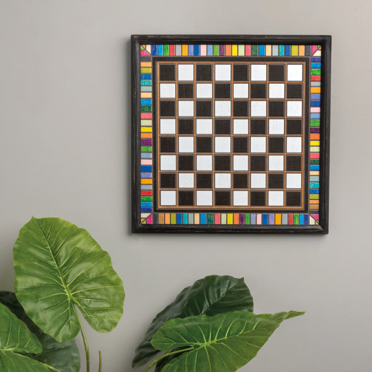 Framed checkered board with colorful border on a gray wall with a plant with green leaves below.
