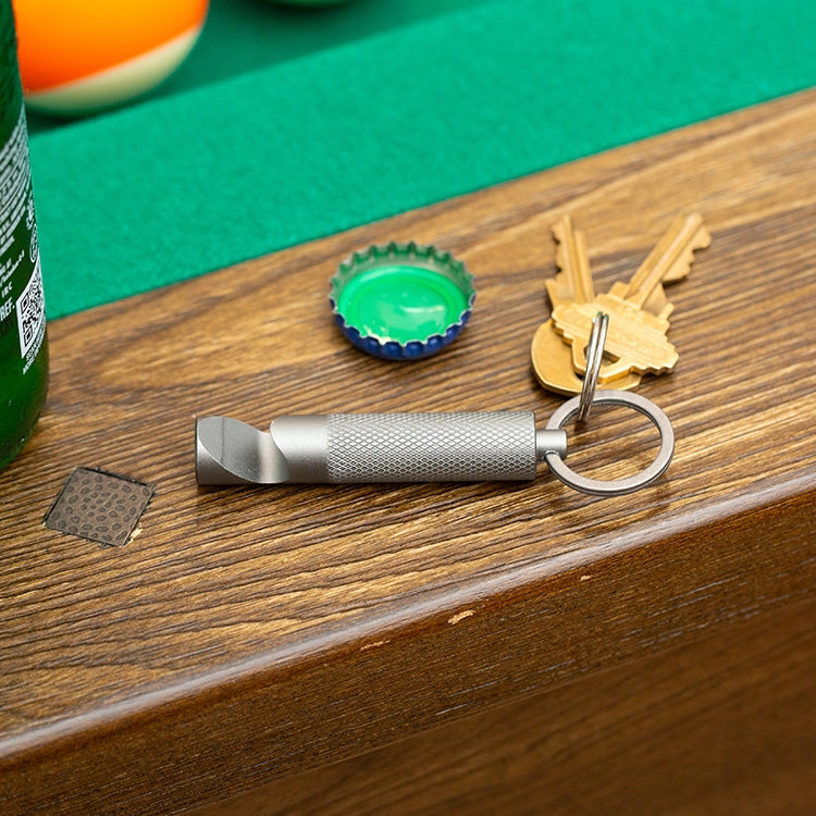 Metal bottle opener on a keychain with keys on a wooden surface next to a bottle cap.