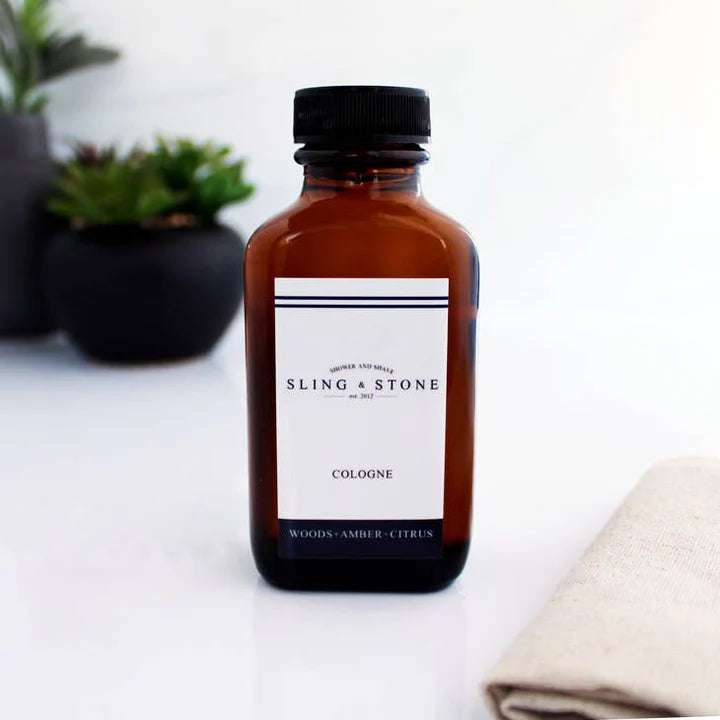 Brown bottle of Sling & Stone cologne on a white surface with plants in the background