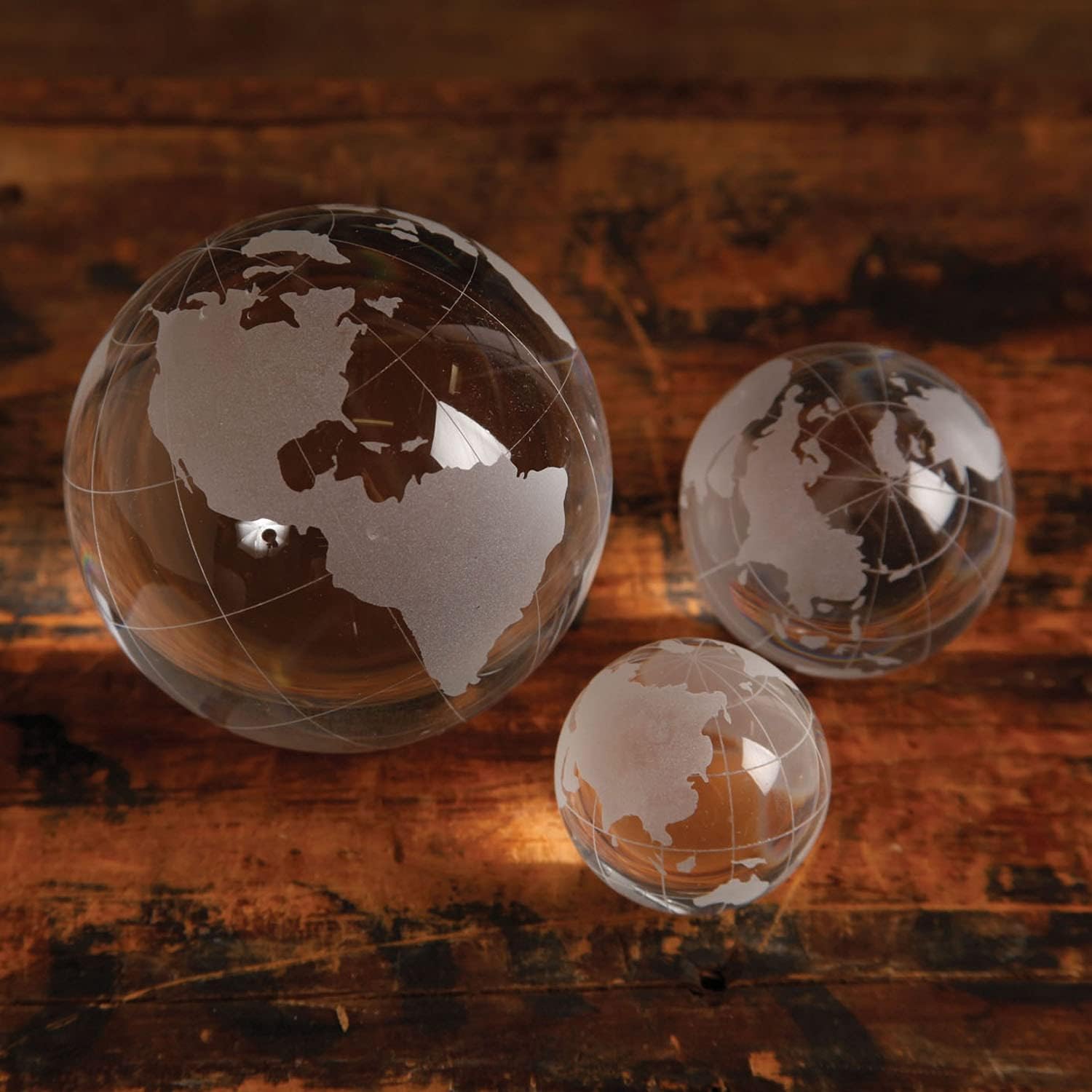 Three etched glass world globe paperweights of different sizes on a wooden surface.