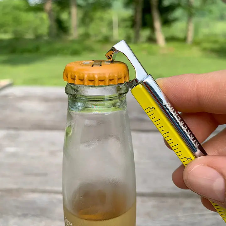 Person using a bottle opener pen a small glass bottle with an orange cap outdoors.