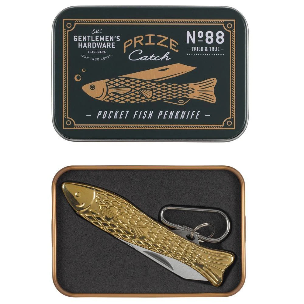 A gold-colored brass pocket fish penknife with 'Gentlemen's Hardware' branding on the blade, displayed in a black and gold tin with a fish-shaped design.