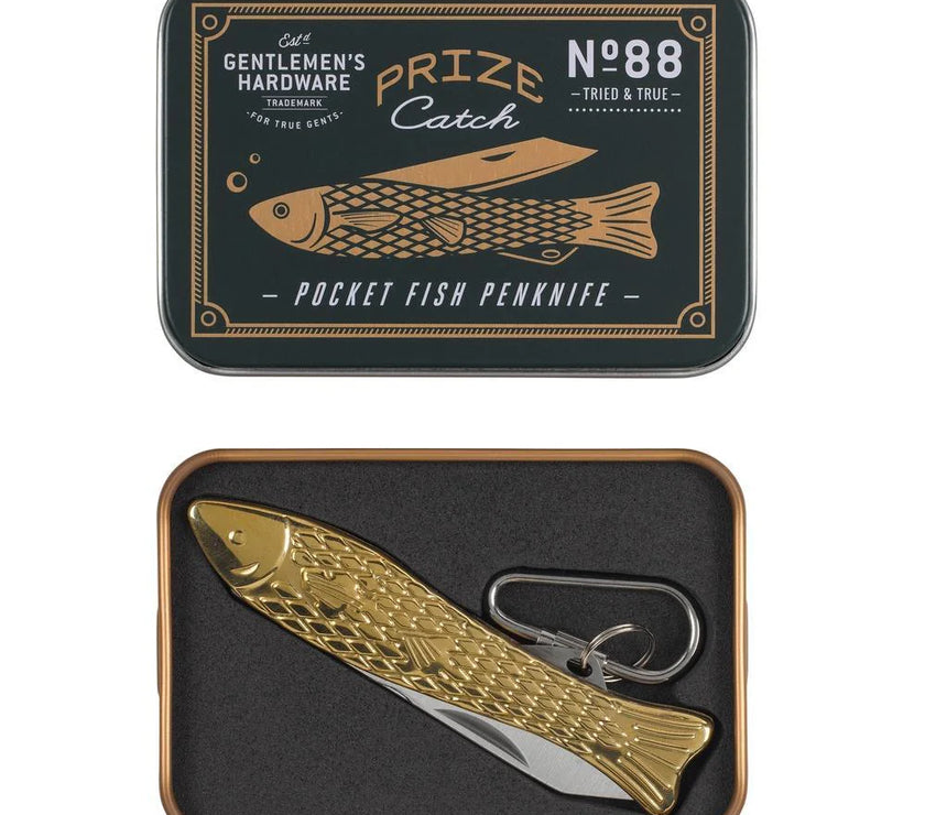 A gold-colored brass pocket fish penknife with 'Gentlemen's Hardware' branding on the blade, displayed in a black and gold tin with a fish-shaped design.