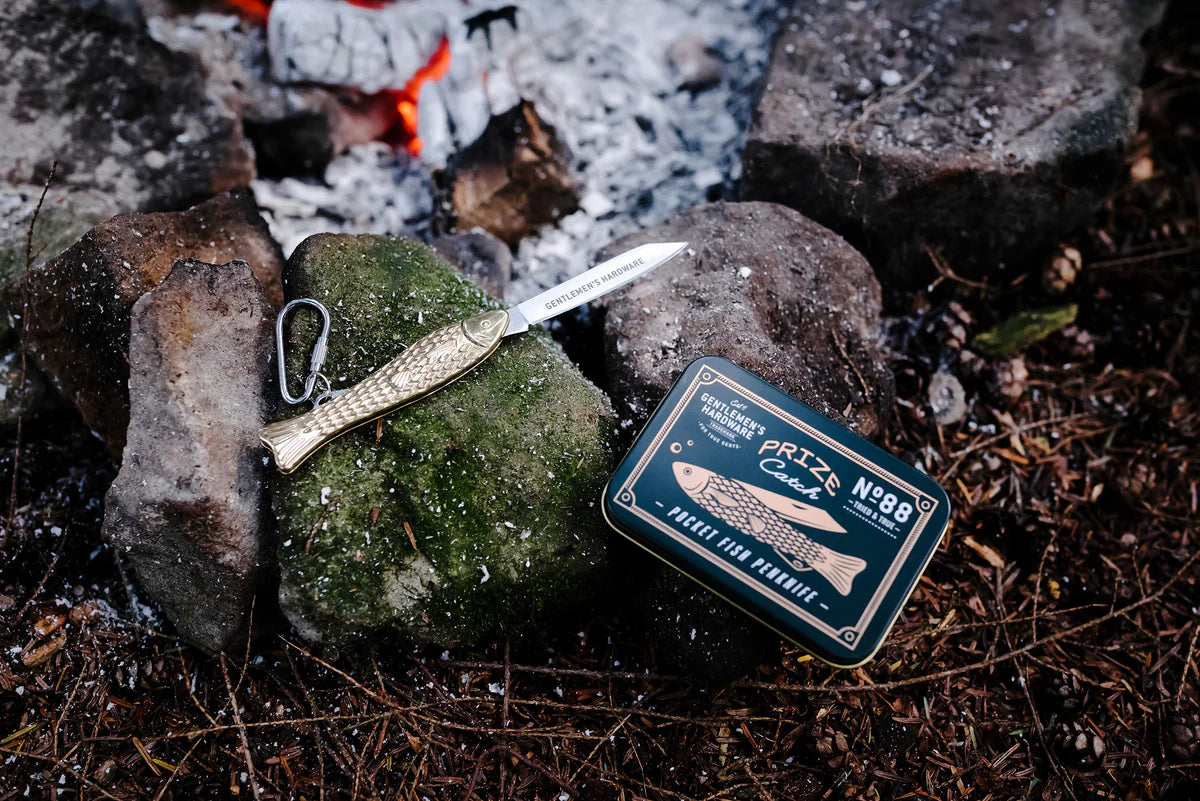 Brass fish pen knife with carabiner and its packaging on rocks near a campfire