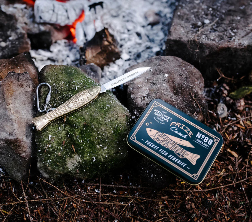 Brass fish pen knife with carabiner and its packaging on rocks near a campfire