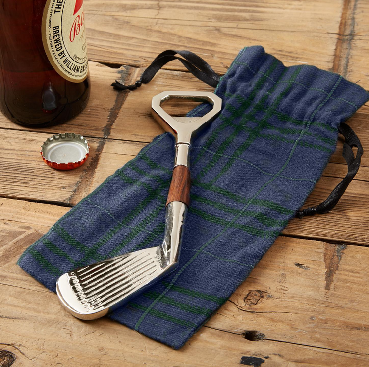 A golf club shaped bottle opener laying on a blue plaid pouch on a wood table with a bottle of beer and bottlecap nearby.