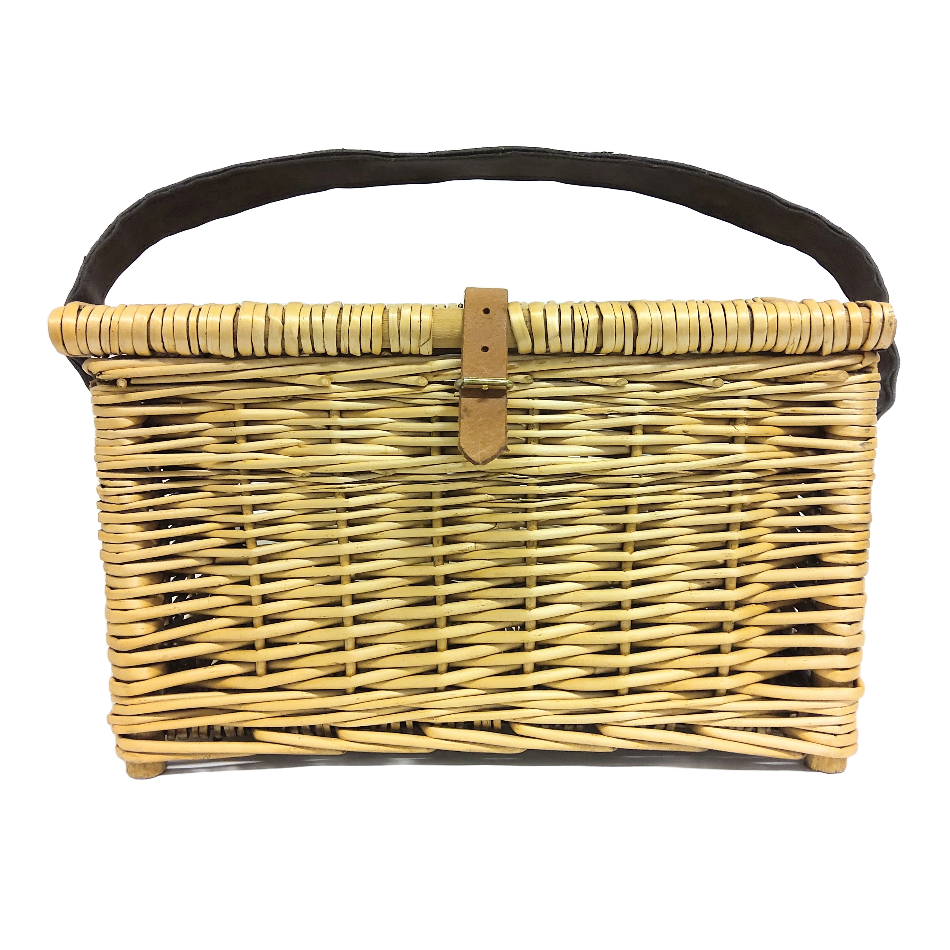 A hand-woven brown fishing basket with a lid and leather carrying strap on a rock near a river with fishing tackle nearby.