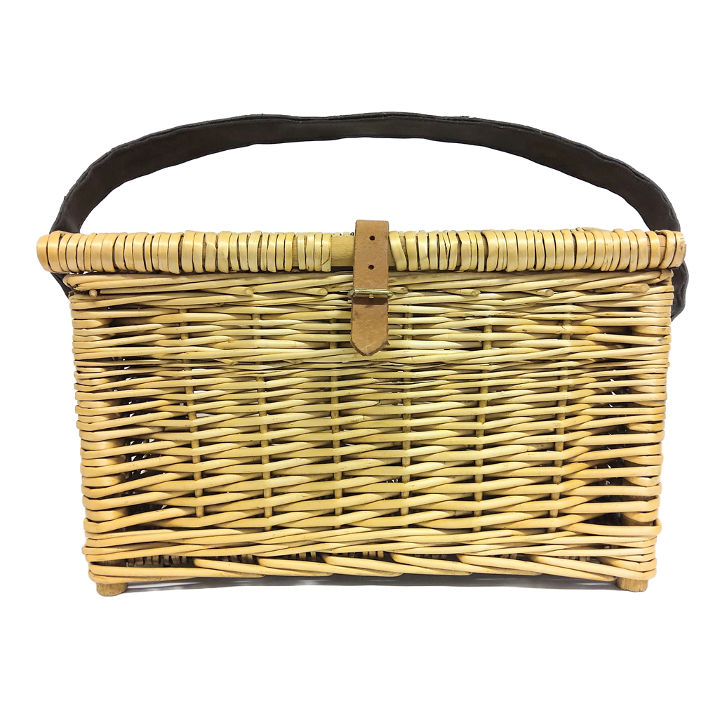 A hand-woven brown fishing basket with a lid and leather carrying strap on a rock near a river with fishing tackle nearby.