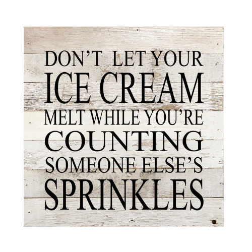 A wooden sign with the phrase, 'DON'T LET YOUR ICE CREAM MELT WHILE YOU'RE COUNTING SOMEONE ELSE'S SPRINKLES' printed in black lettering against a white background.