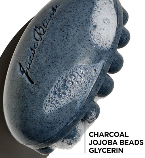 A closeup of a gray Jack Black Charcoal Body Bar with massaging nubs, with text pointing out the charcoal, jojoba beads, and glycerin.
