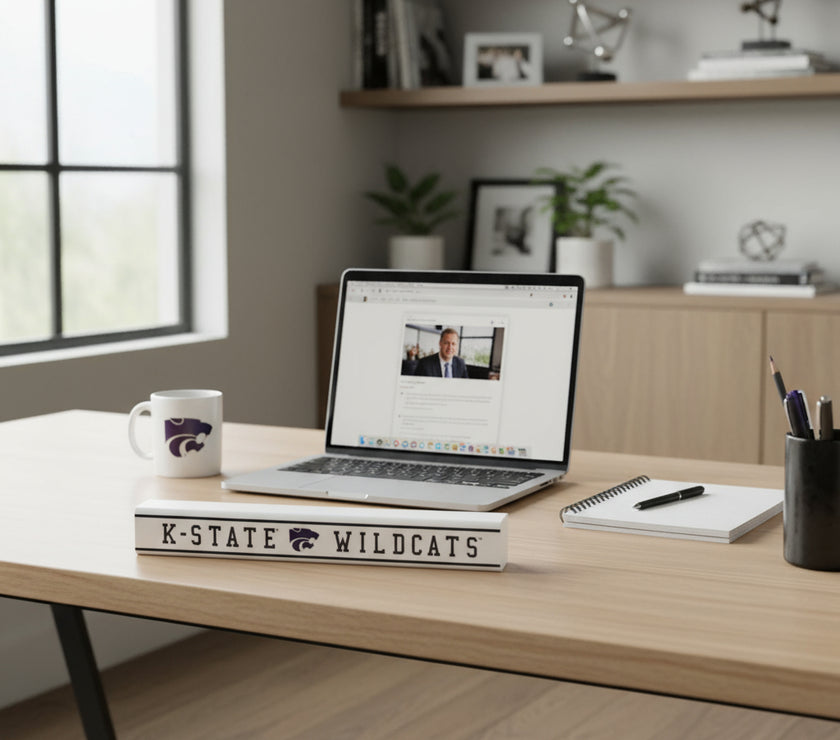 A rectangular pine wood sign with 'K-State Wildcats' text, placed on a desk with a computer and coffee mug and other office items in the background.