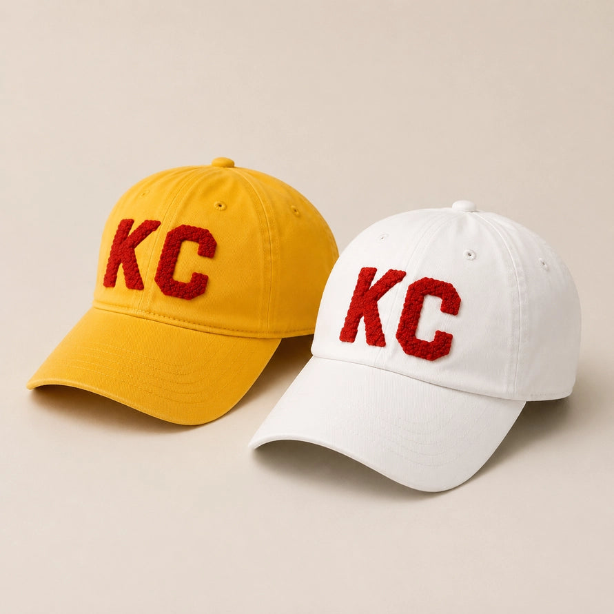 Two baseball caps on a warm beige background: one yellow and one white, each with large red embroidered 'KC' lettering on the front.