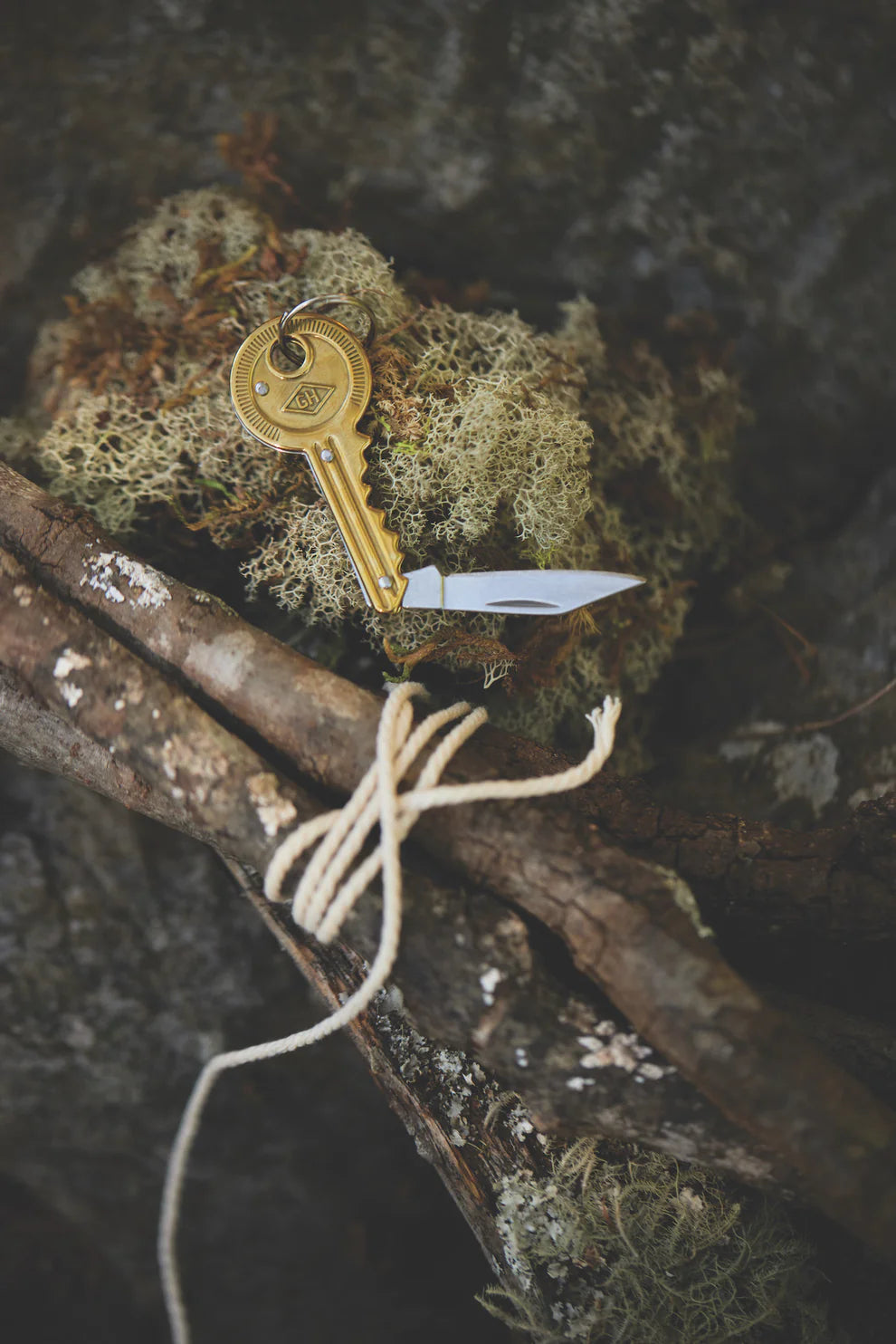 A key-shaped pocket knife lying on a piece of moss next to a bundle of sticks wrapped with string.
