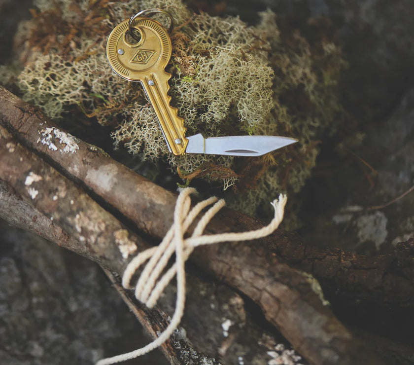A key-shaped pocket knife lying on a piece of moss next to a bundle of sticks wrapped with string.