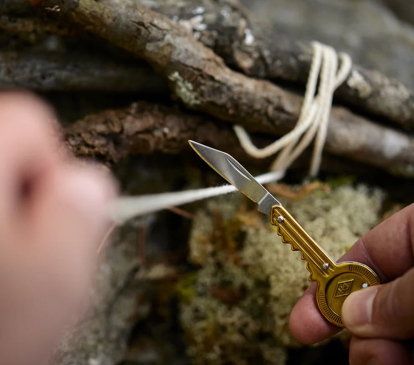 Hand holding a key with a pocket knife attached against a piece of string with a natural background.