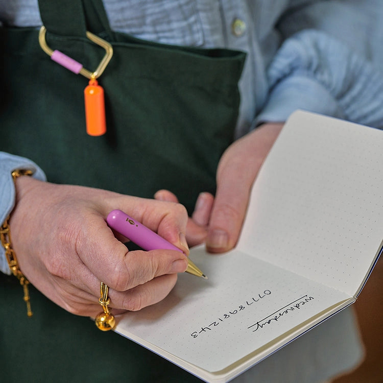 Person writing in a notebook with a pink pen, wearing a green bag with colorful handles.