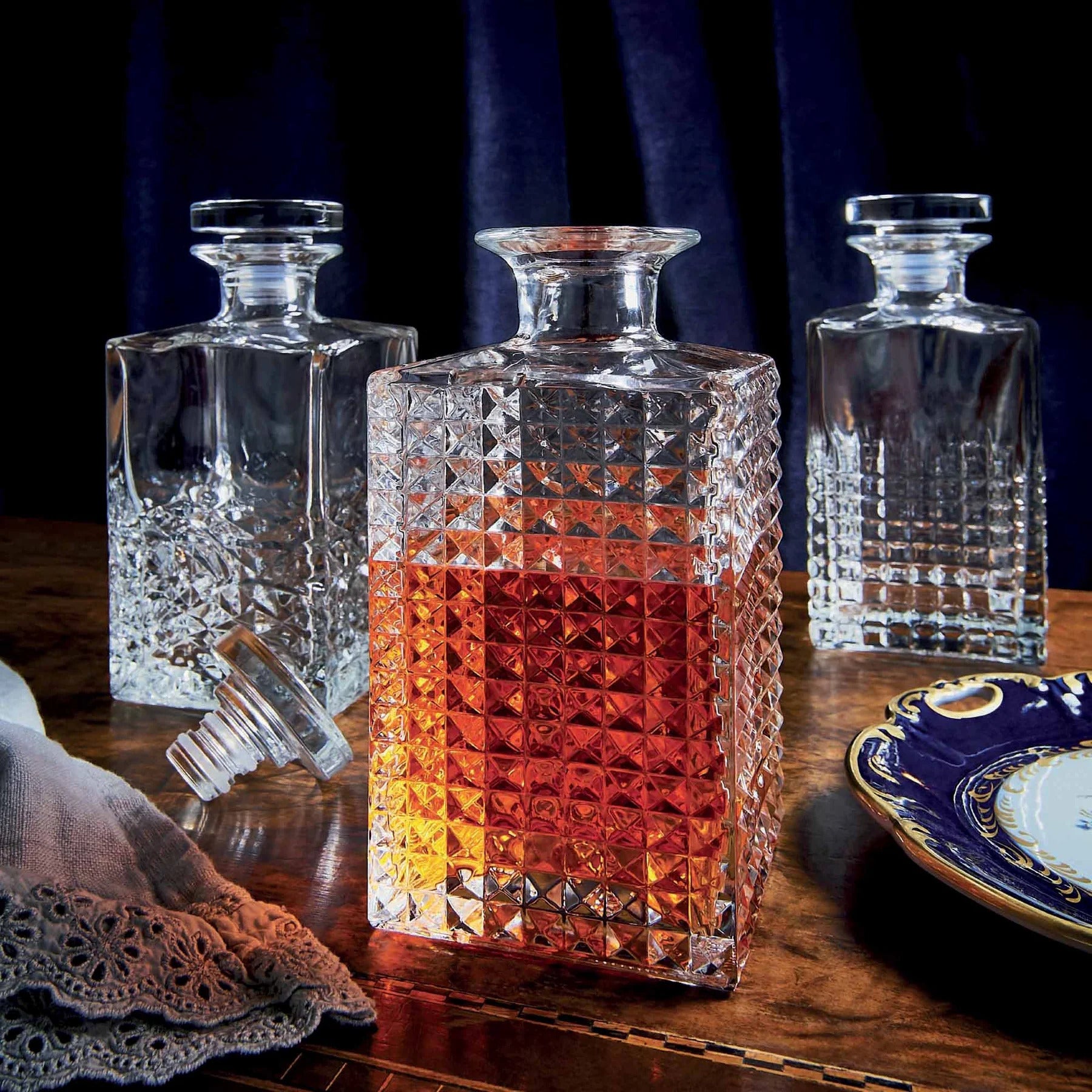 A clear glass decanter with a geometric pattern design with liquor in it on a table with identical decanters.