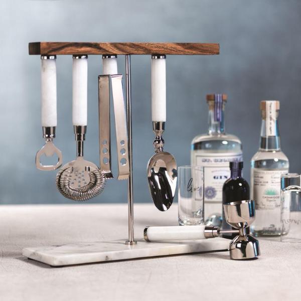 A five piece bar tool set on a white surface with bottles and drinking glasses nearby with a gray background.