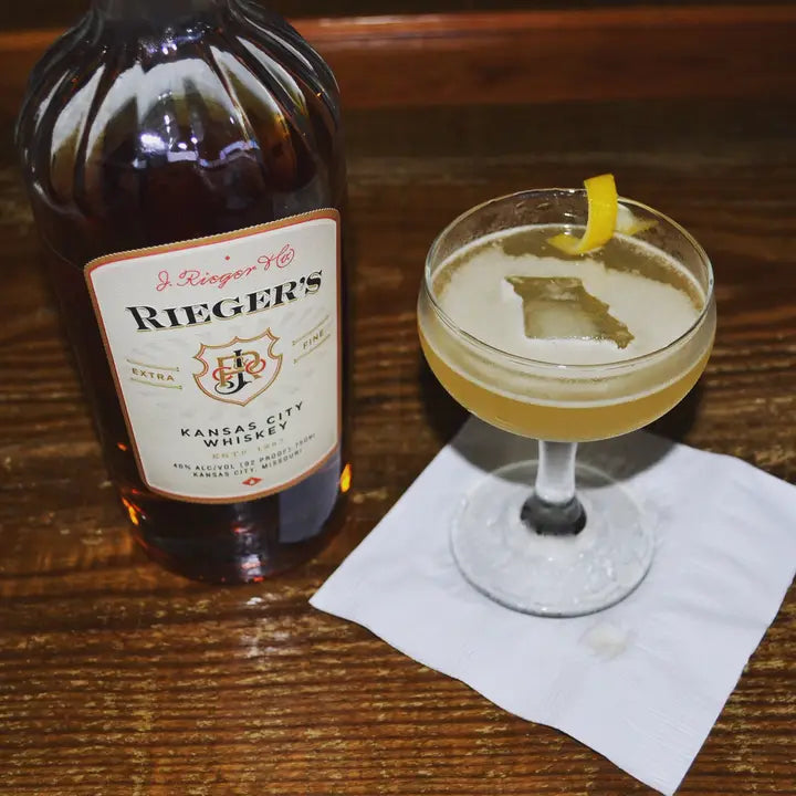 A cocktail with a single ice cube in the shape of the state of Missouri in it with a bottle of Rieger's next to it.