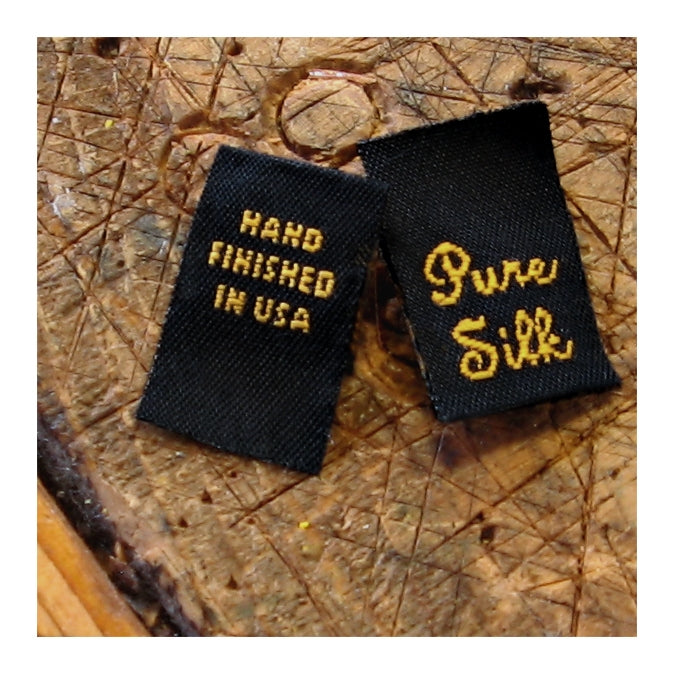 Two black pouches with words stitched on in gold thread on a wooden surface, one says 'hand finished in usa' and the other says 'pure silk.'