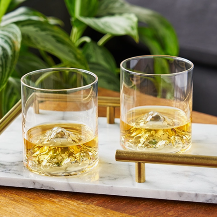Two glass tumblers with mountain shapes crafted in the bottoms, filled with liquid, sit on a marble tray with a plant in the background.