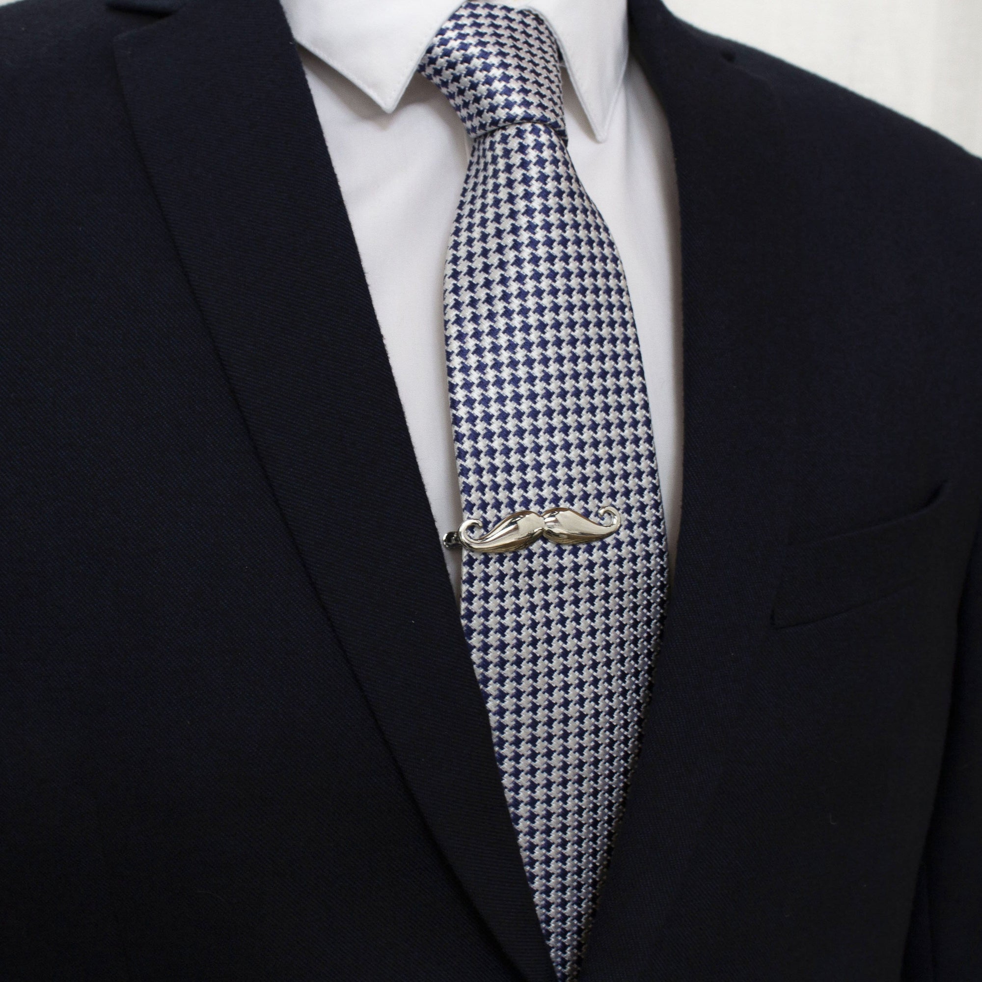 A silver mustache tie bar clipped to a blue and white houndstooth patterned tie worn by a person in a suit.
