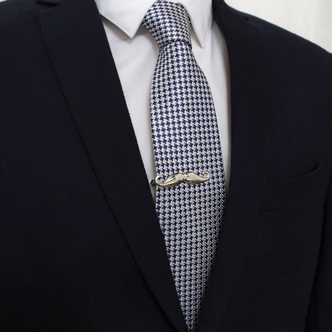 A silver mustache tie bar clipped to a blue and white houndstooth patterned tie worn by a person in a suit.