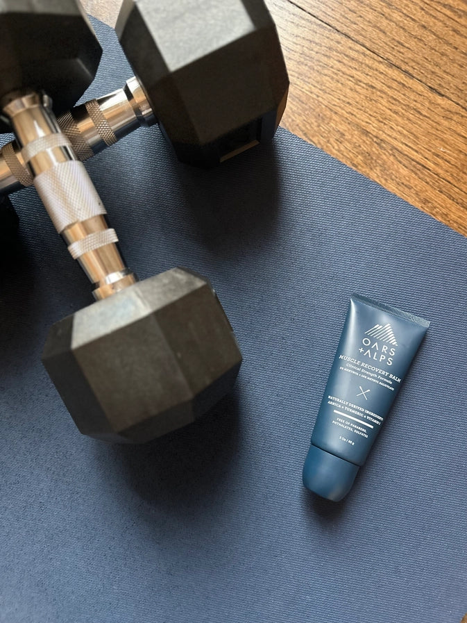 A tube of Oars + Alps Muscle Recovery Balm lying on a workout mat next to a pair of dumbells. 