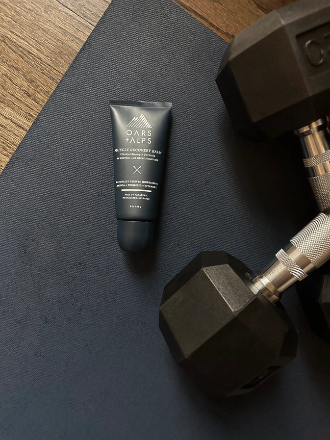 A tube of Oars + Alps Muscle Recovery Balm lying on a workout mat next to a pair of dumbells. 