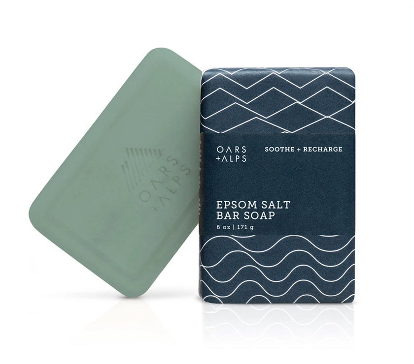A bar of soap with a greenish tint next to its packaging, which has a blue and white pattern. The packaging reads 'Epsom Salt Bar Soap'.