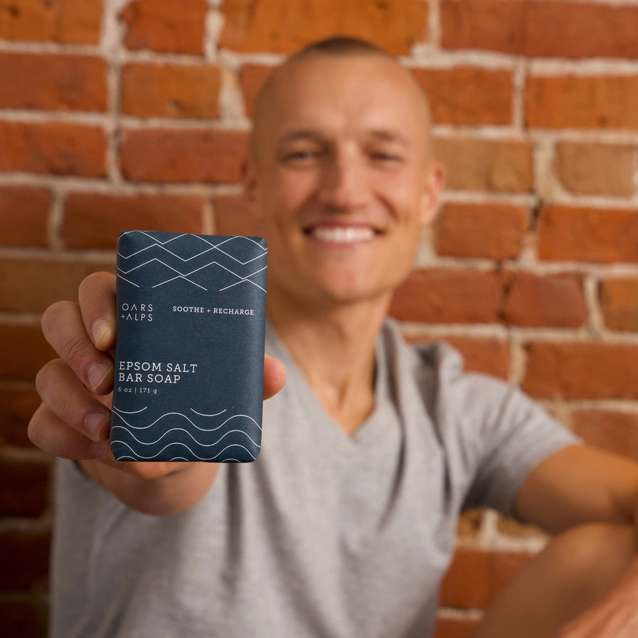 A person holding Oars and Alps Epsom Salt Bar Soap wrapped in its dark blue paper package.