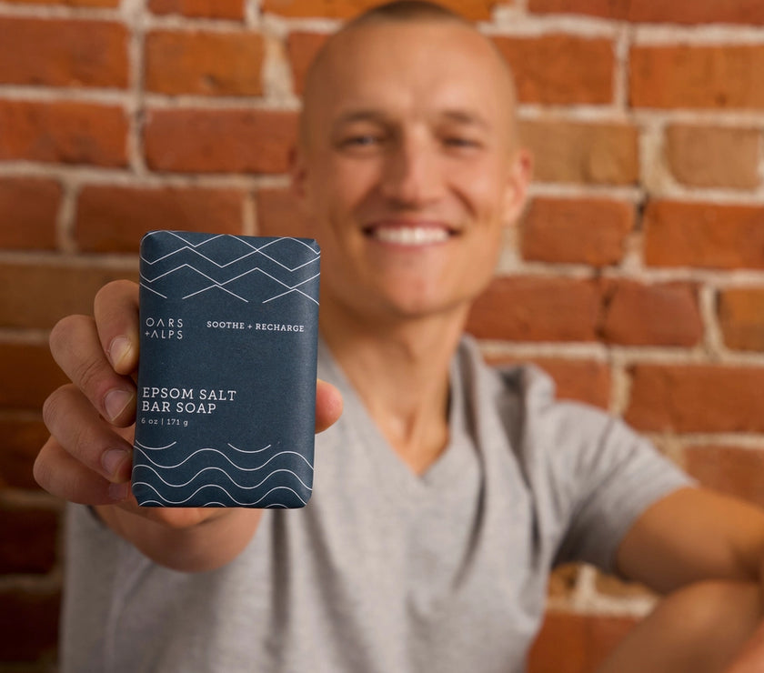 A person holding Oars and Alps Epsom Salt Bar Soap wrapped in its dark blue paper package.