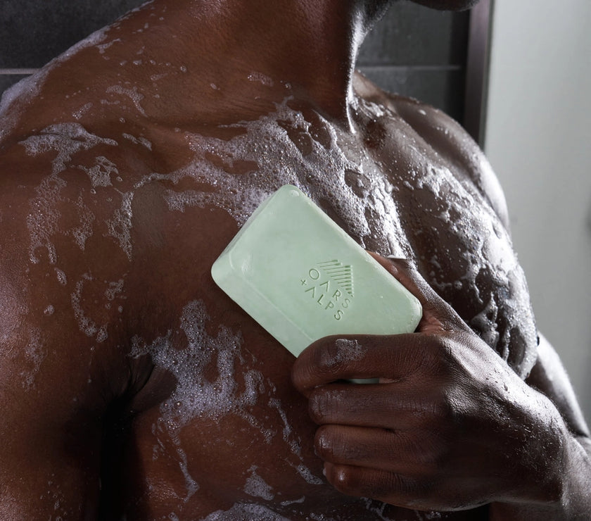 A man holding a bar of Oars and Alps Epsom Salt soap against his chest in the shower.
