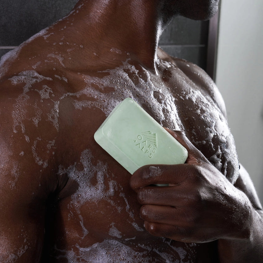 A man holding a bar of Oars and Alps Epsom Salt soap against his chest in the shower.