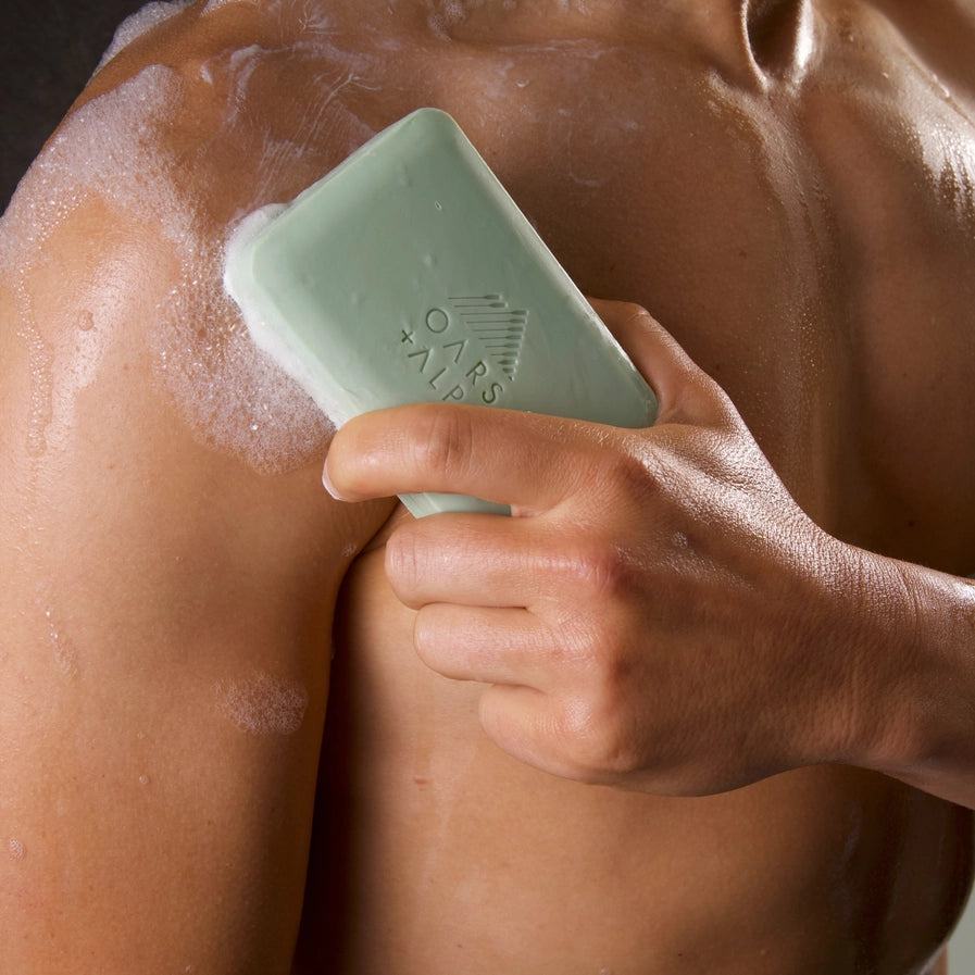 A man holding a bar of Oars and Alps Epsom Salt soap against his chest in the shower.