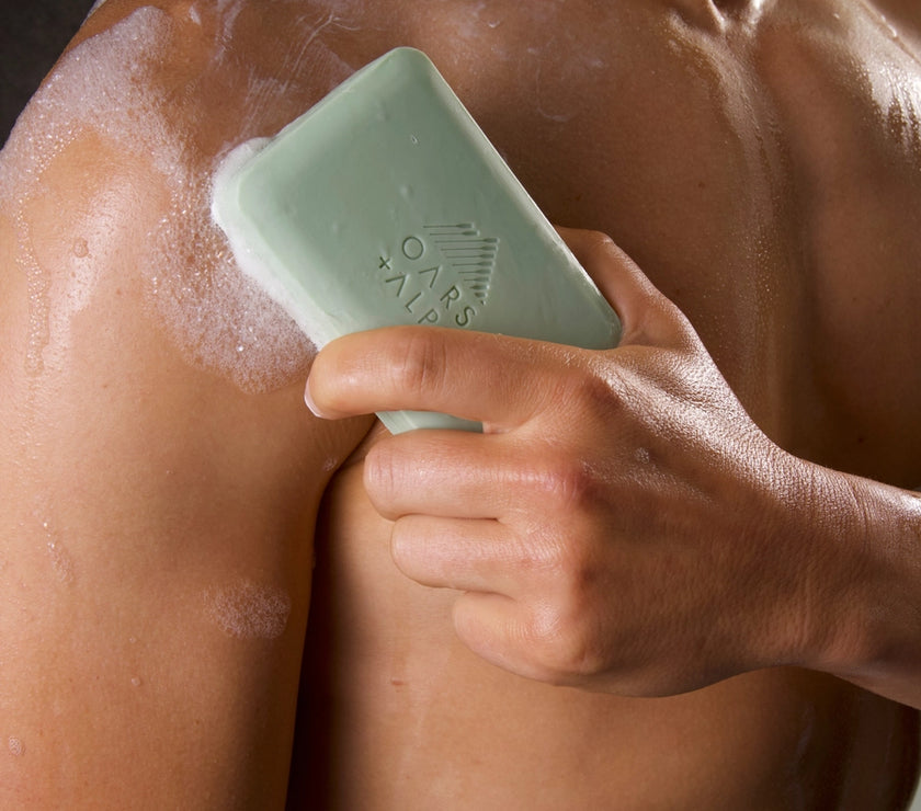 A man holding a bar of Oars and Alps Epsom Salt soap against his chest in the shower.