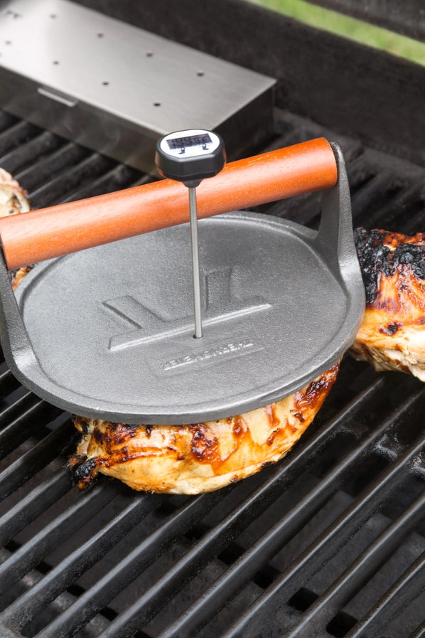 Black meat thermometer inserted through a hole in a cast iron grill press and into a piece of chicken on a grill.