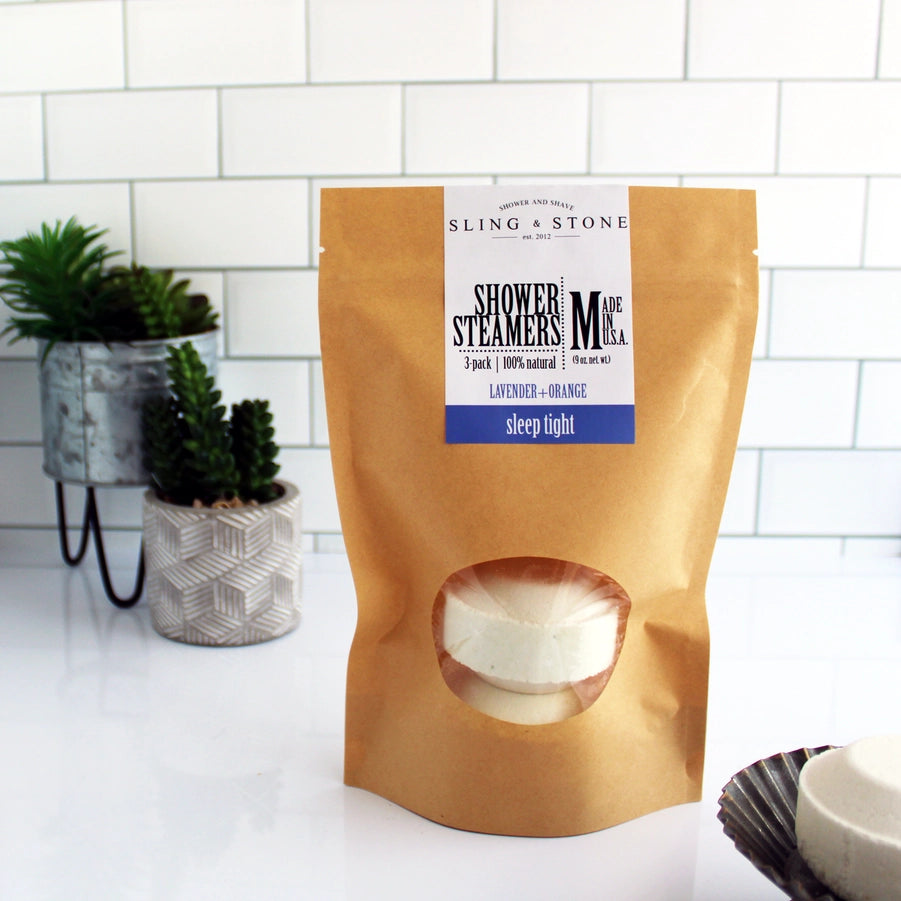 A package of Sling & Stone shower steamers with three visible white tablets through a craft peek-a-boo window bag, placed on a white countertop with a plant in the background.