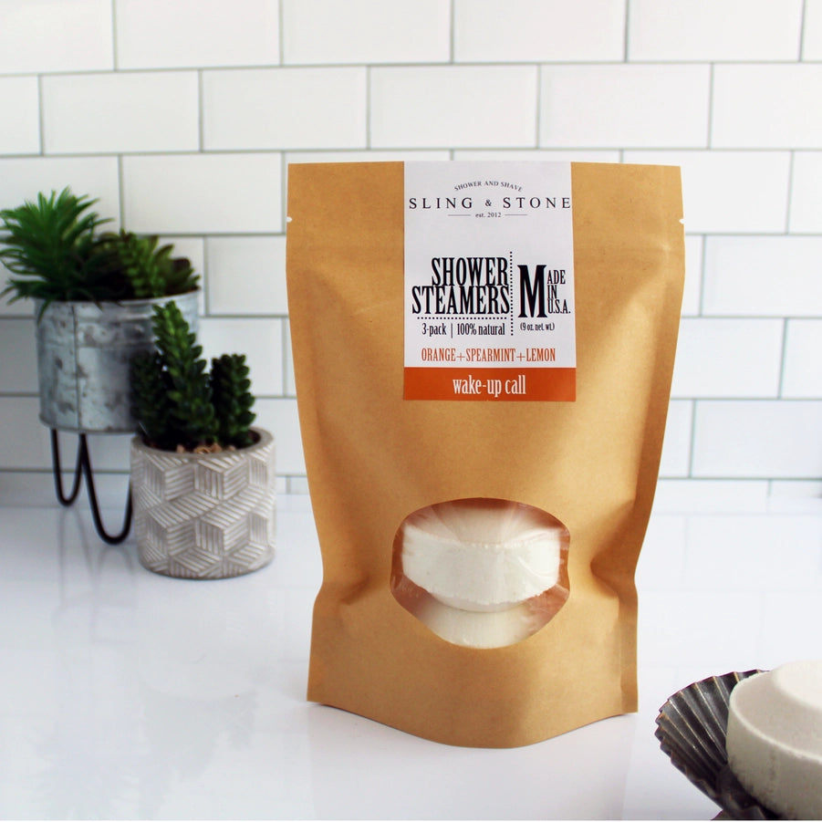 A package of Sling & Stone shower steamers with three visible white tablets through a craft peek-a-boo window bag, placed on a white countertop with a plant in the background.