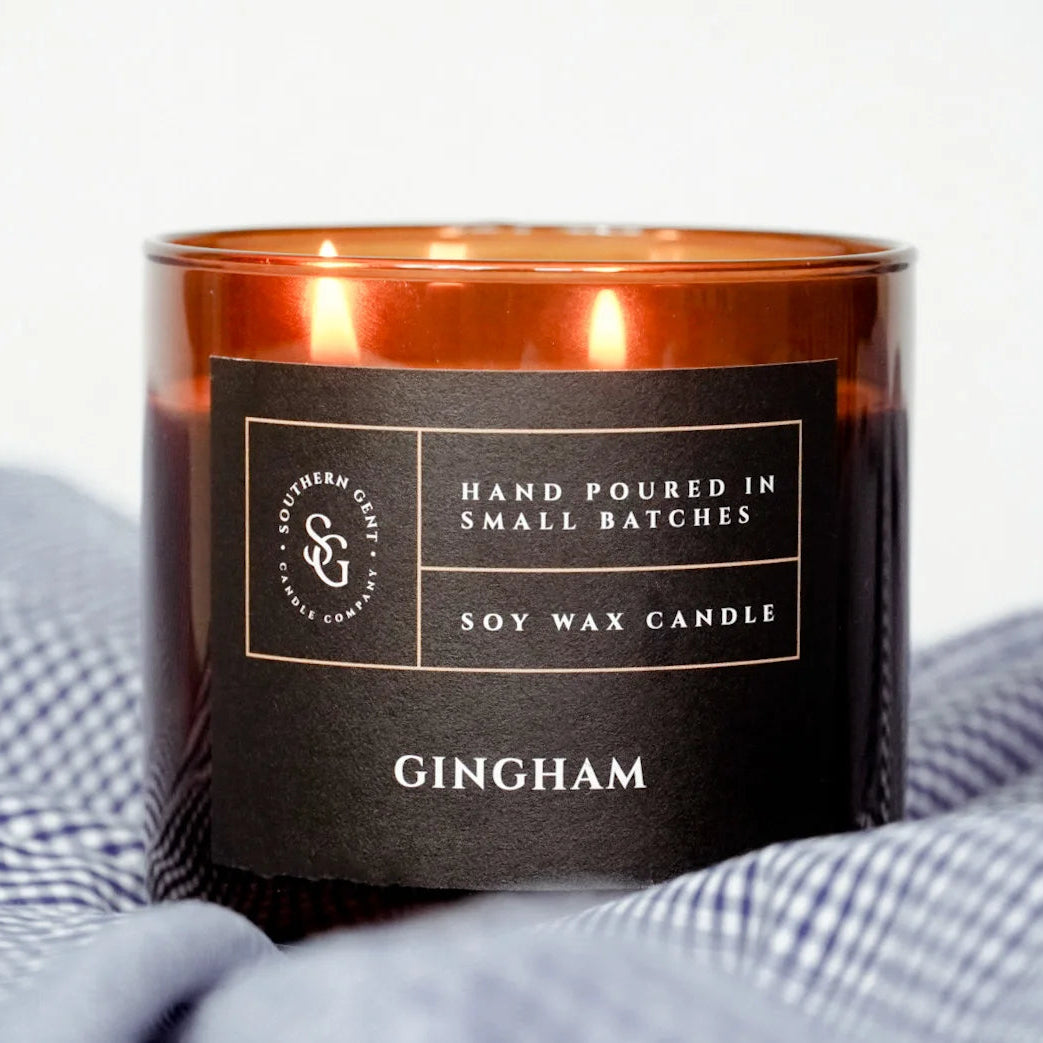 A two-wick candle with a gingham pattern design, placed on a checkered fabric, with a black label that reads 'Gingham' and 'HAND POURED IN SMALL BATCHES SOY WAX CANDLES'.

