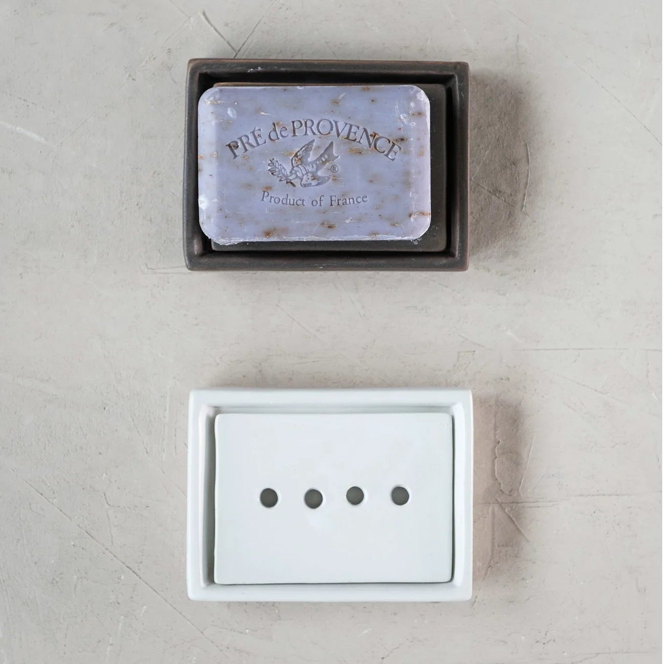 White and brown stoneware soap dishes with a bar of soap on the brown on on a textured neutral background.