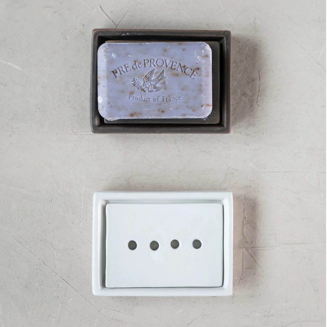 White and brown stoneware soap dishes with a bar of soap on the brown on on a textured neutral background.