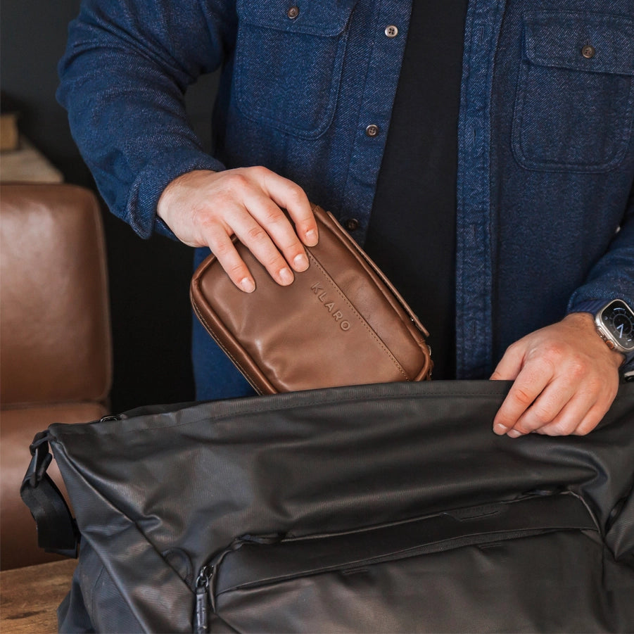A man in a blue jacket putting a brown leather travel cigar case into a black bag.