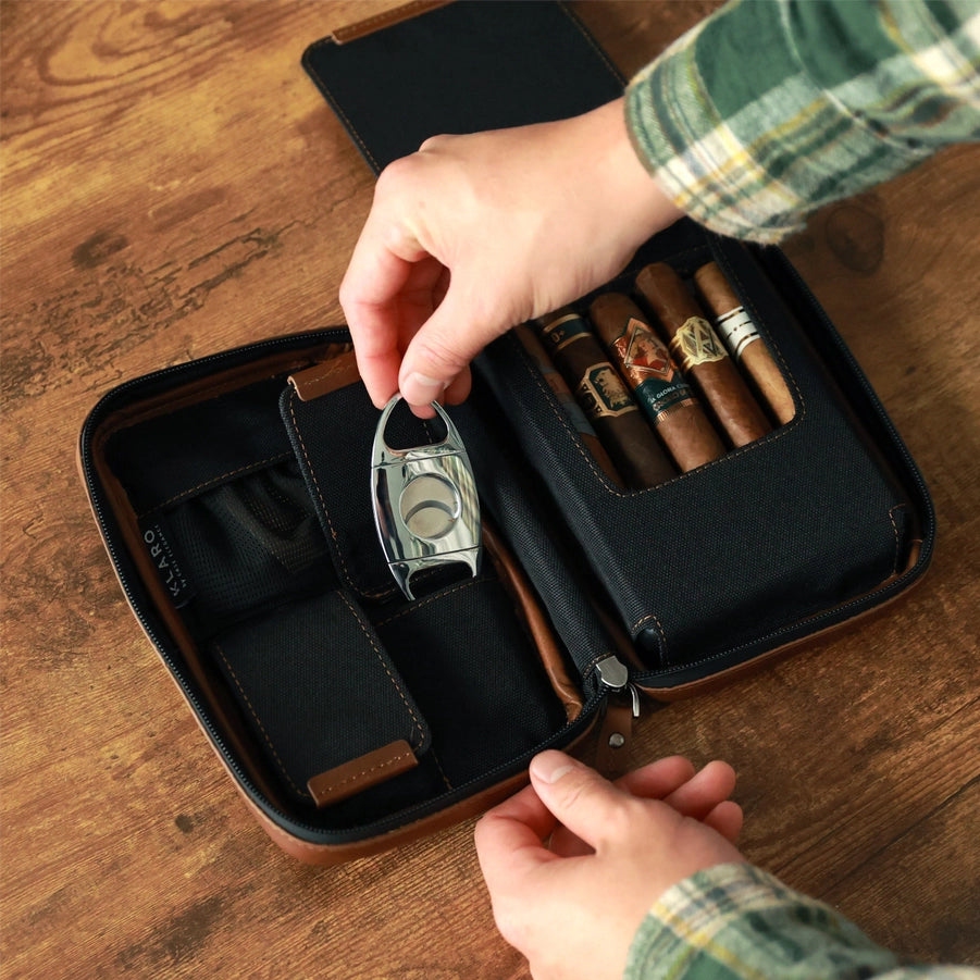 A person using a brown leather travel cigar case that includes a black interior and compartments for cigars and cigar accessories on a wooden surface.