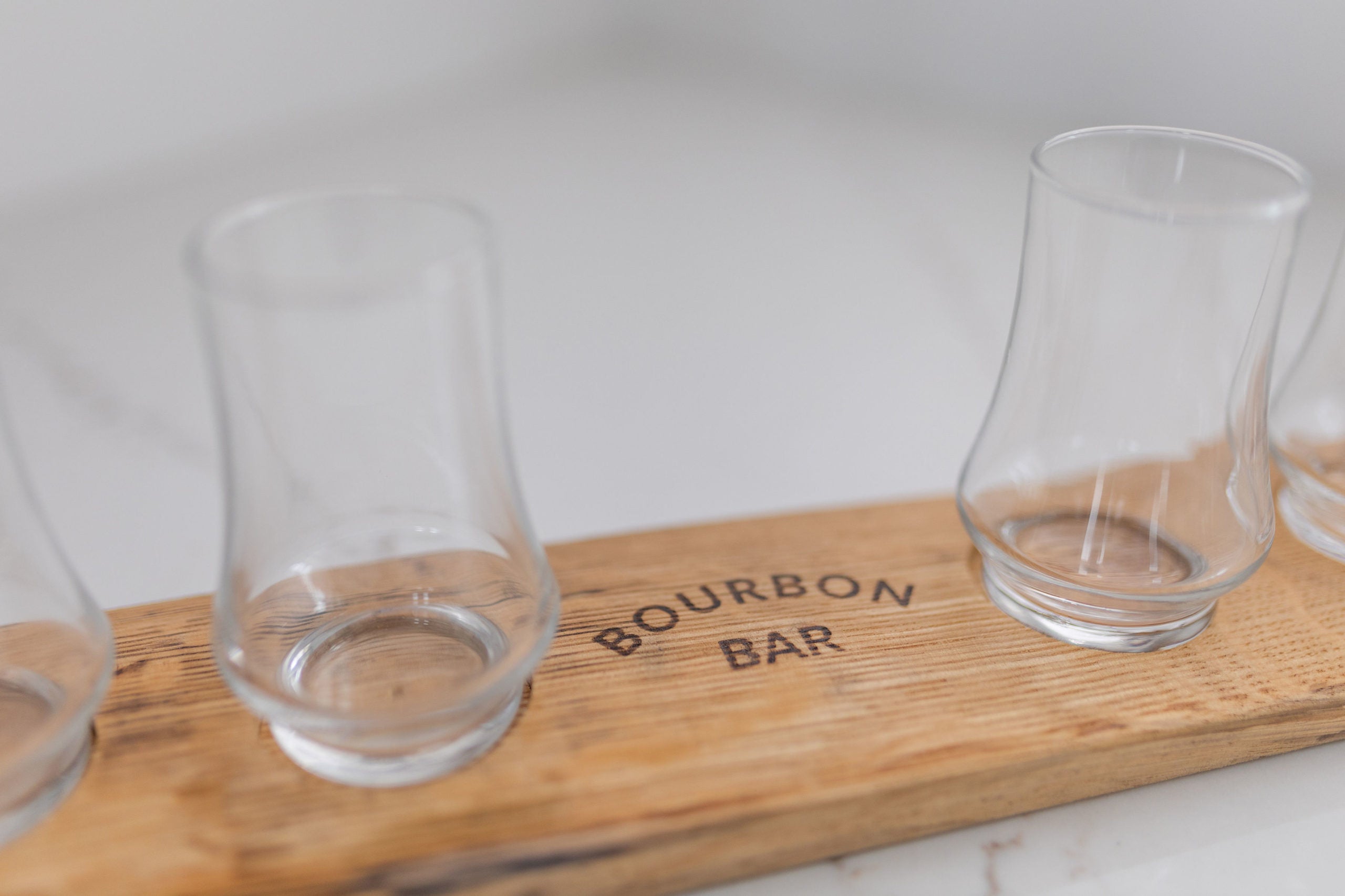 A wooden tasting flight board with four clear whiskey glasses and 'BOURBON BAR' on it, placed on a marble surface.