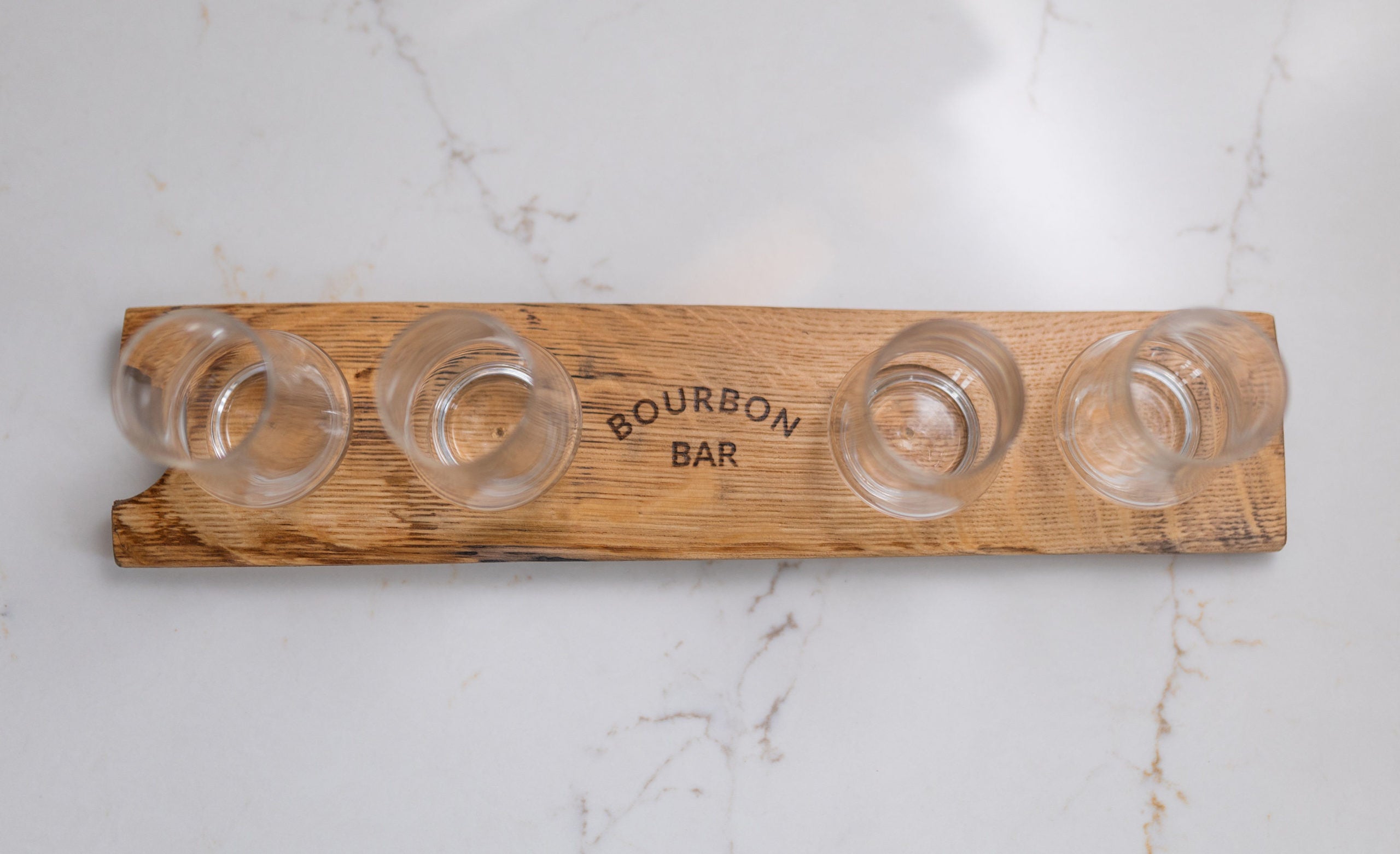 A wooden tasting flight board with four clear whiskey glasses and 'BOURBON BAR' on it, placed on a marble surface.