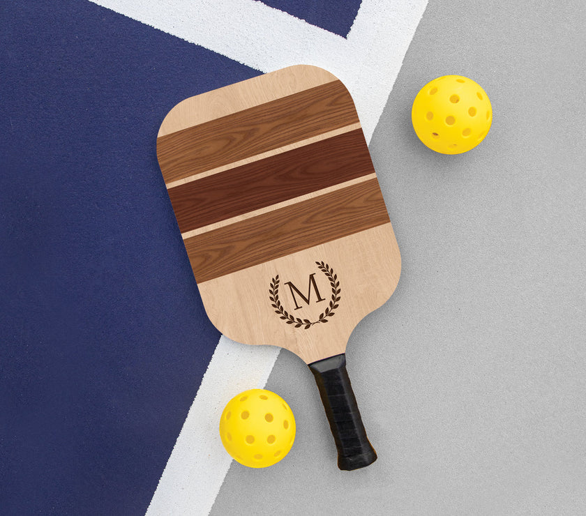 Customizable wooden pickleball paddle with a natural wood finish, displaying the text 'M' on the center and a small logo below it. The paddle is placed on a grey surface with two yellow pickleballs to the side.