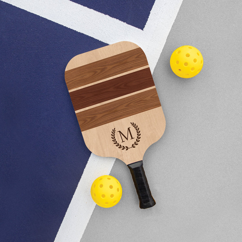 Customizable wooden pickleball paddle with a natural wood finish, displaying the text 'M' on the center and a small logo below it. The paddle is placed on a grey surface with two yellow pickleballs to the side.