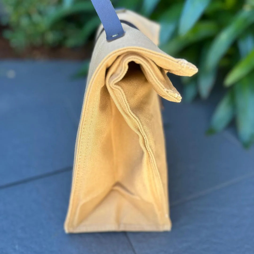 Side view of a yellow canvas lunch bag with a black strap and buckle-fastening closure on a dark gray surface with blurred plants in the background.