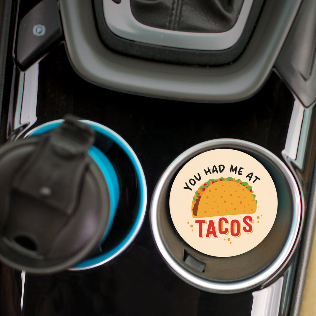 A circular car coaster with 'you had me at tacos' written on it with an image of a taco inside of a car cup holder.